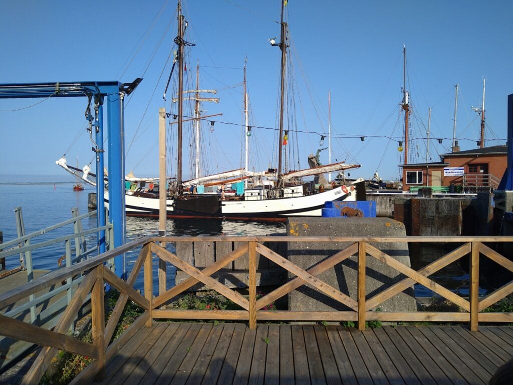 Traditionelles Segelschiff mit hohen Masten liegt im sonnigen Hafen; Holzsteg und Geländer im Vordergrund.