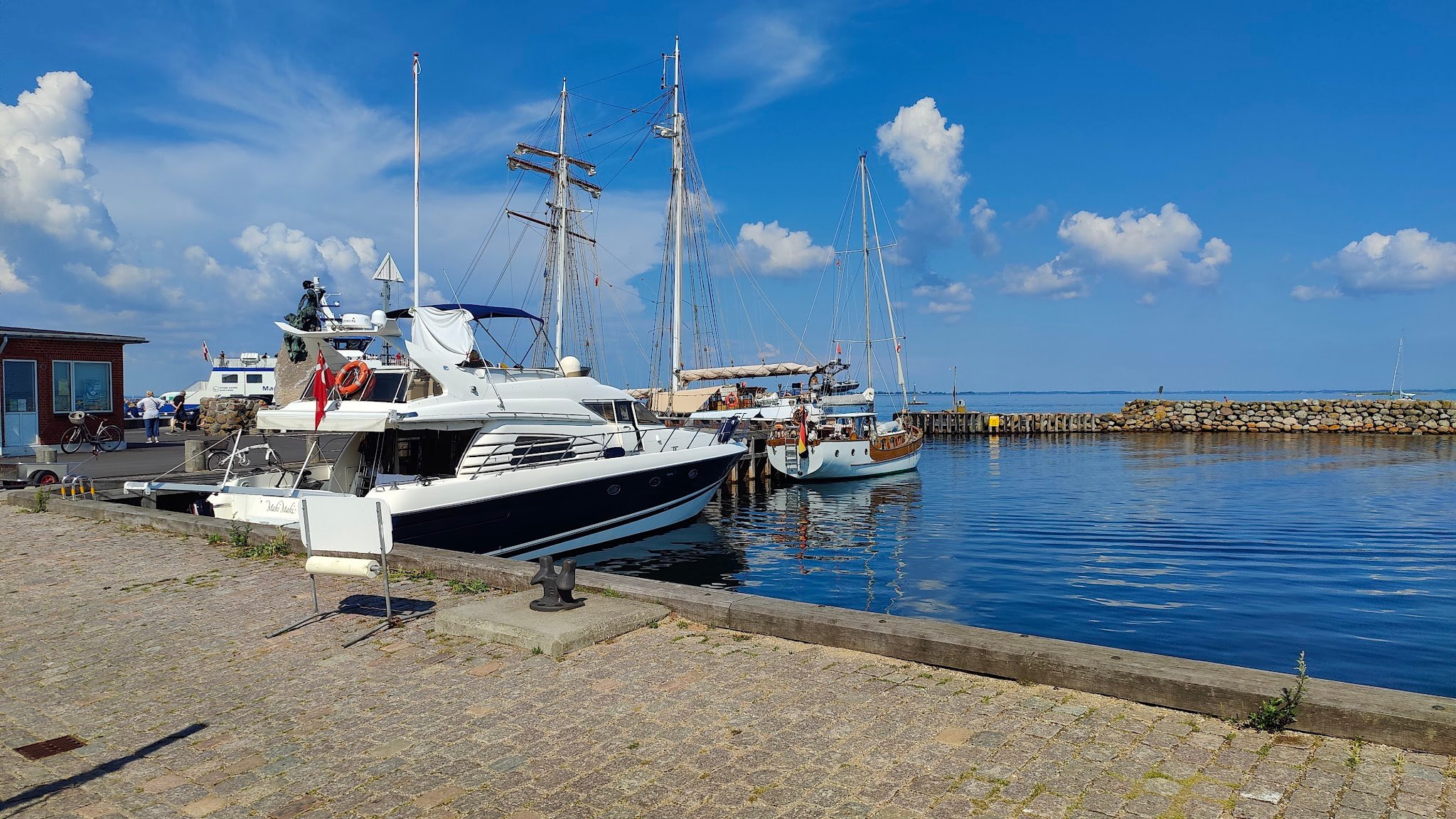Motorjacht und mehrere Segelboote liegen in ruhigem Hafen; blauer Himmel, vereinzelte Wolken.