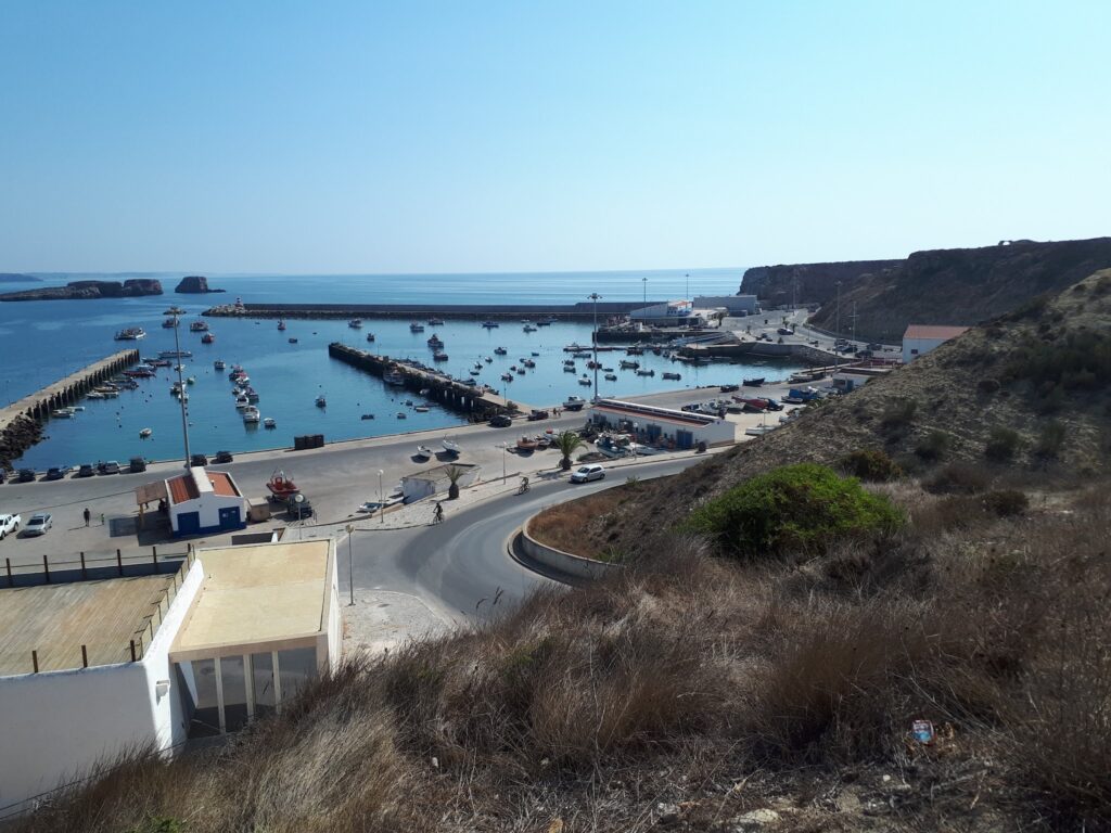 Kleine Boote im geschützten Hafen zwischen zwei Piers, umgeben von Klippen und blauem Meer unter klarem Himmel.