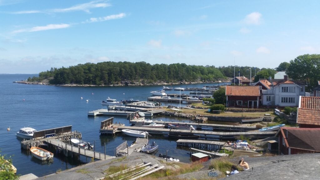 Sonniger schwedischer Küstenhafen: rote Holzhäuser, Stege mit Booten, bewaldete Inseln im ruhigen Wasser.