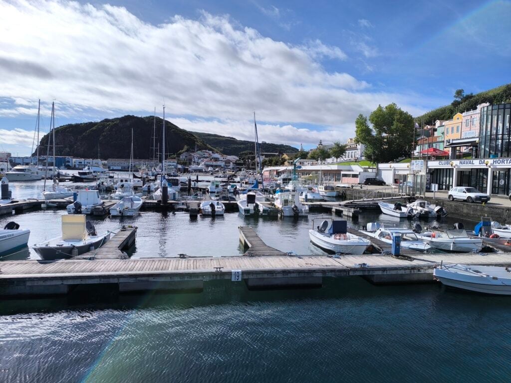Kleiner Hafen mit anliegenden Motor- und Segelbooten, dahinter Hügel und bunte Häuser unter blauem Himmel.