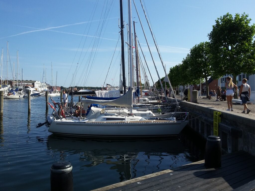 Segelboote im sonnigen Hafen; Spaziergänger schlendern am Kai entlang.