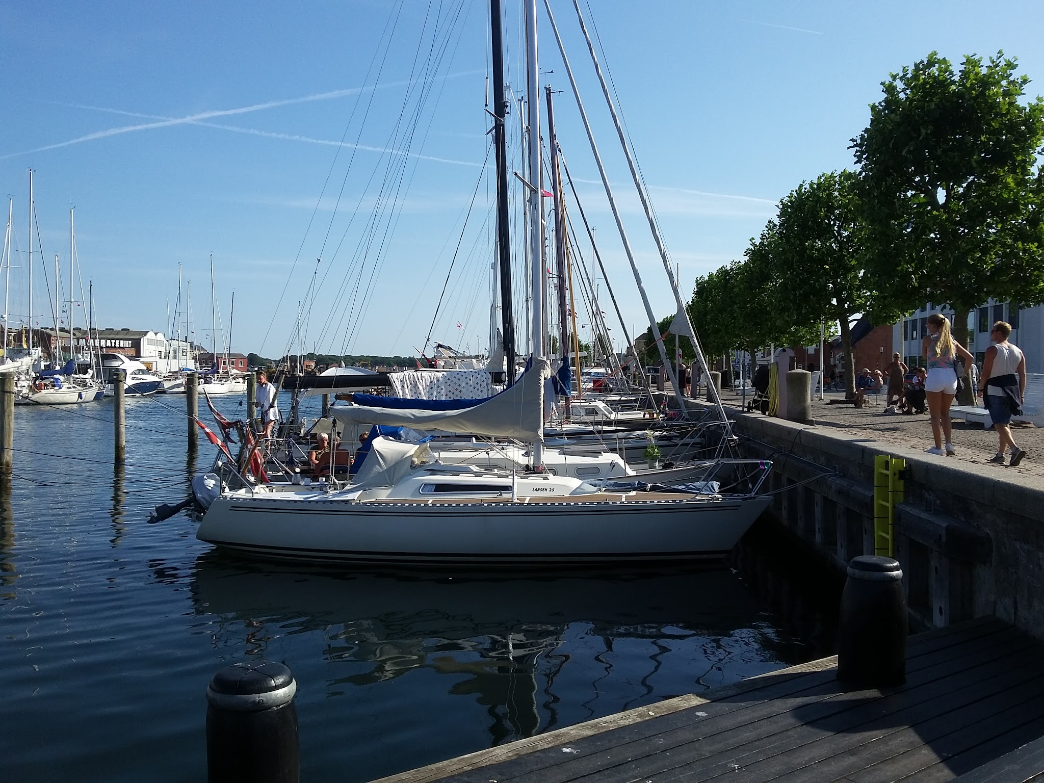 Segelboote im sonnigen Hafen; Spaziergänger schlendern am Kai entlang.