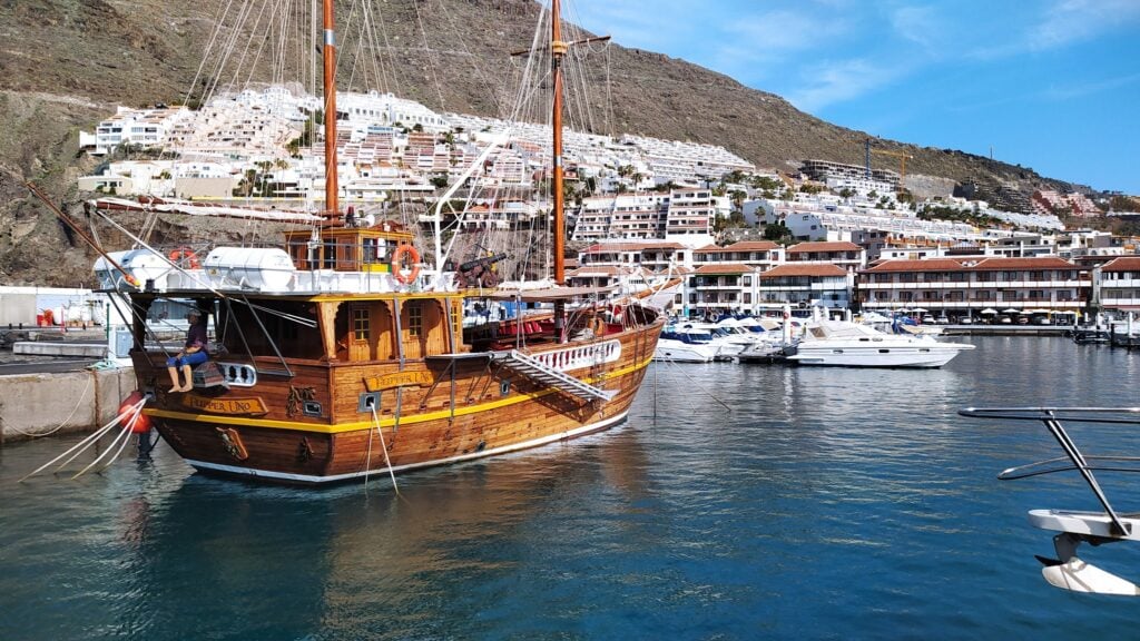 Historisches Holzsegelschiff im sonnigen Yachthafen; weiße Hanghäuser spiegeln sich im Wasser.
