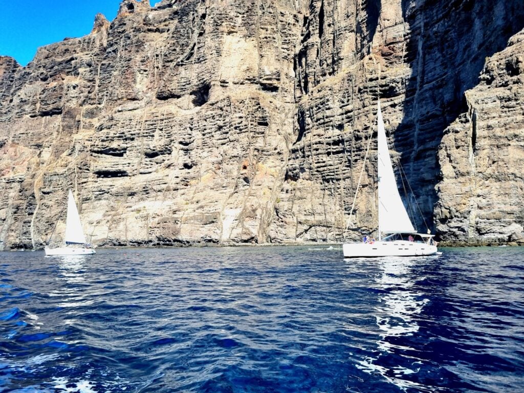 Zwei weiße Segelboote gleiten vor steiler, sonnenbeschienener Felsklippe über tiefblaues Meer.