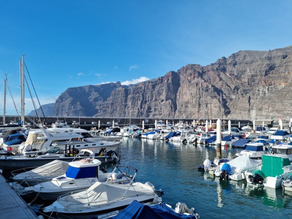 Yachthafen mit zahlreichen weißen Booten im klaren Wasser; dahinter hohe, felsige Klippen unter blauem Himmel