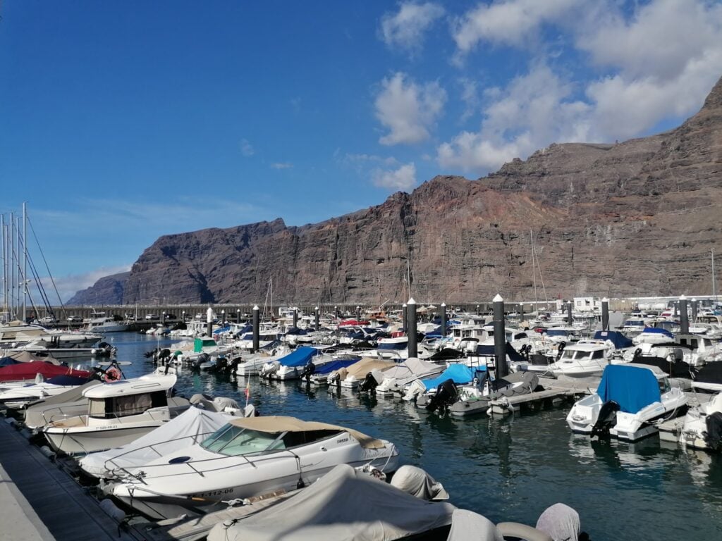 Voller Yachthafen vor hohen, braunen Steilklippen unter klarem, blauem Himmel.