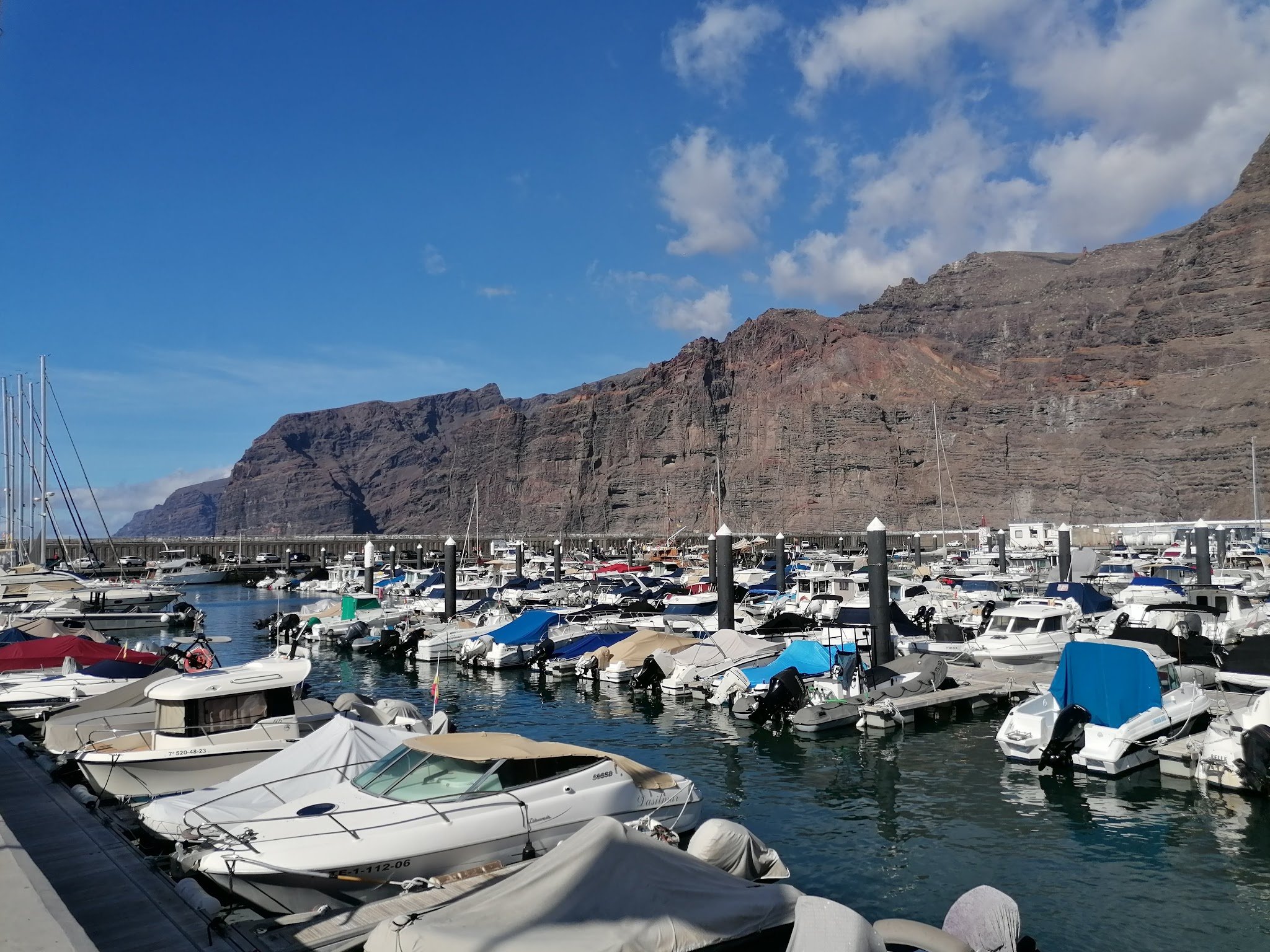 Voller Yachthafen vor hohen, braunen Steilklippen unter klarem, blauem Himmel.
