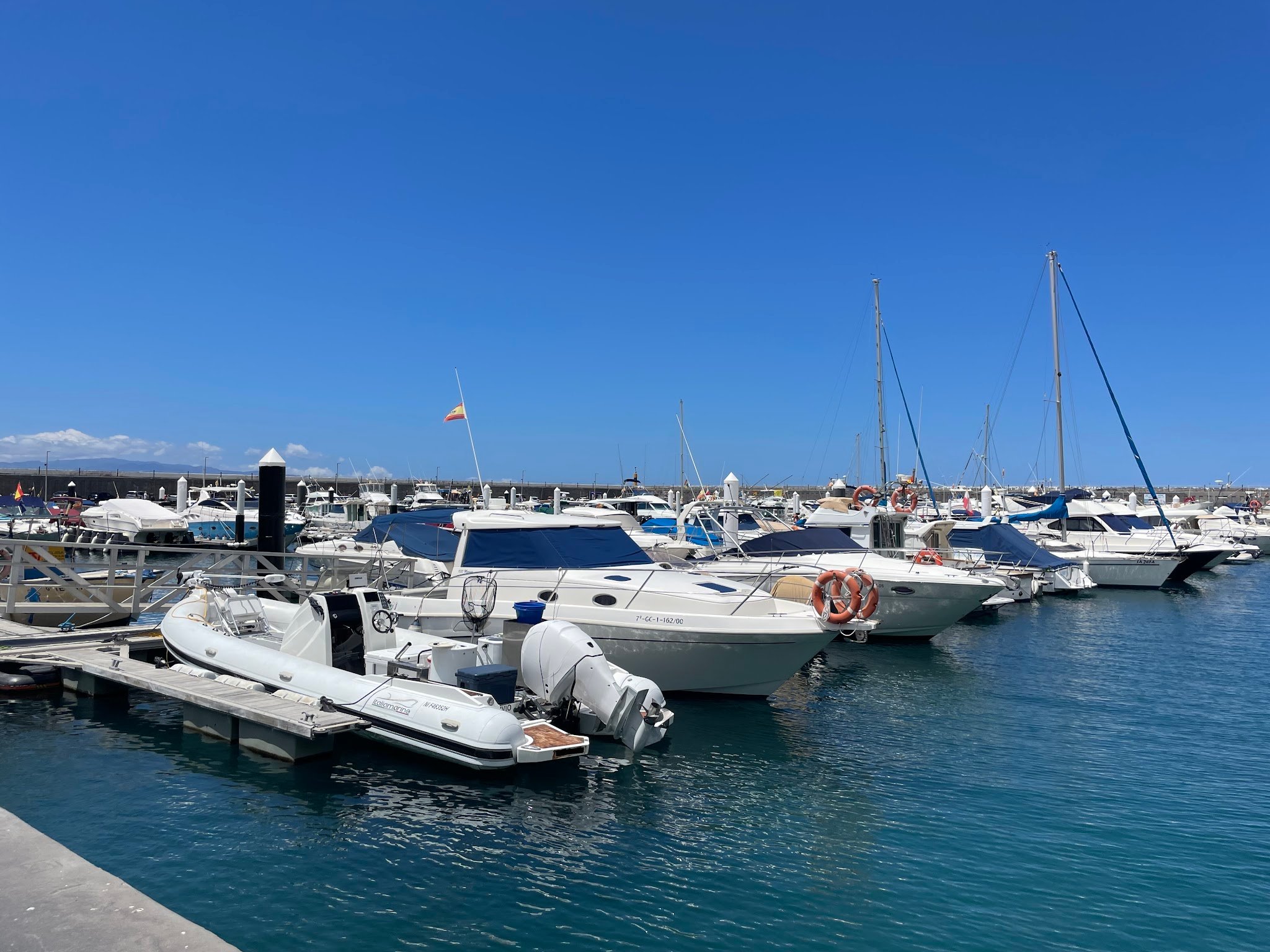Reihe weißer Motorboote und Yachten im sonnigen Hafen, ruhiges türkisblaues Wasser, klarer Himmel, spanische Flagge weht.