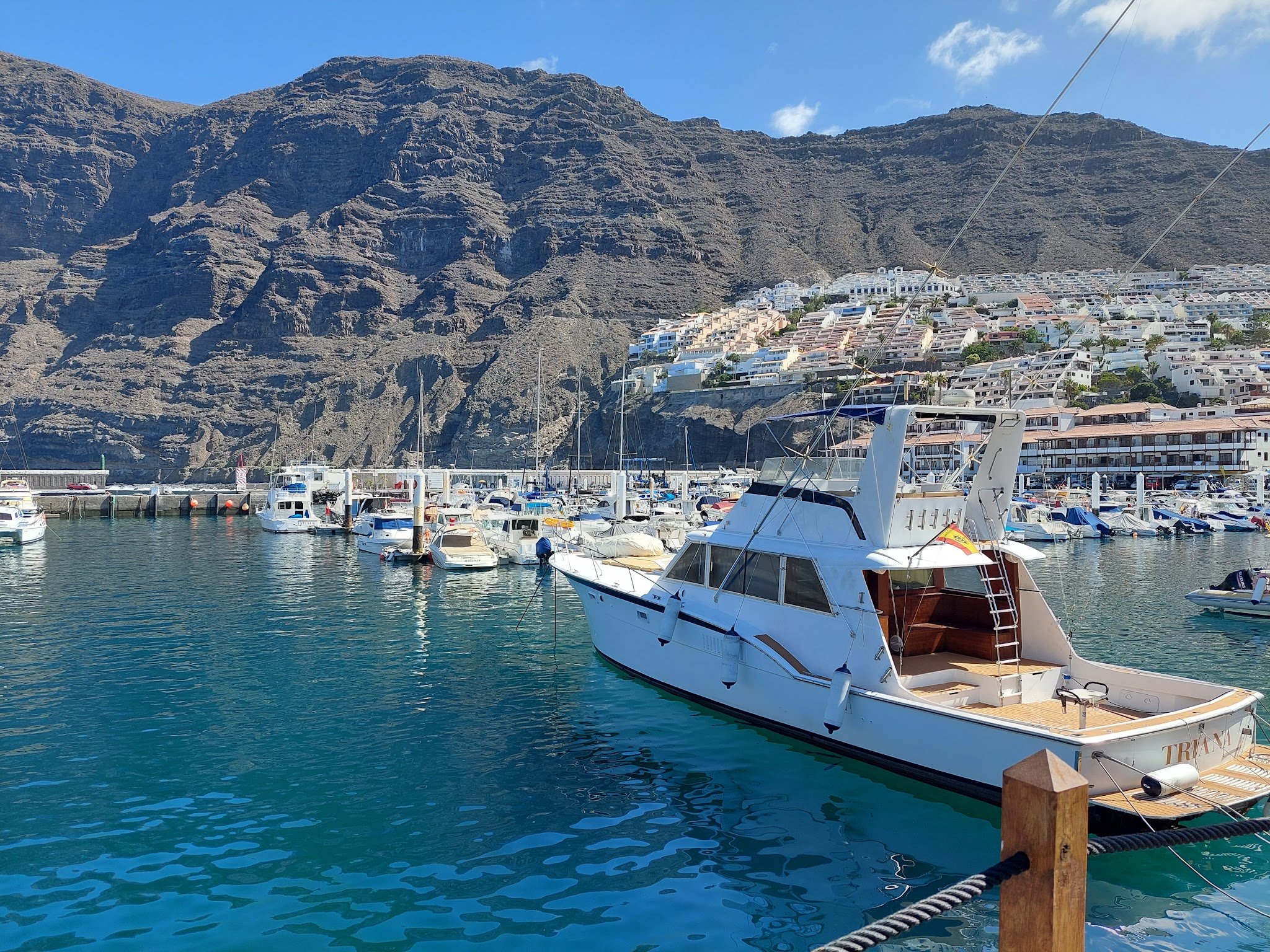 Marina mit weißer Yacht auf tiefblauem Wasser; steile Felswand und terrassenförmig bebaute Küstensiedlung dahinter