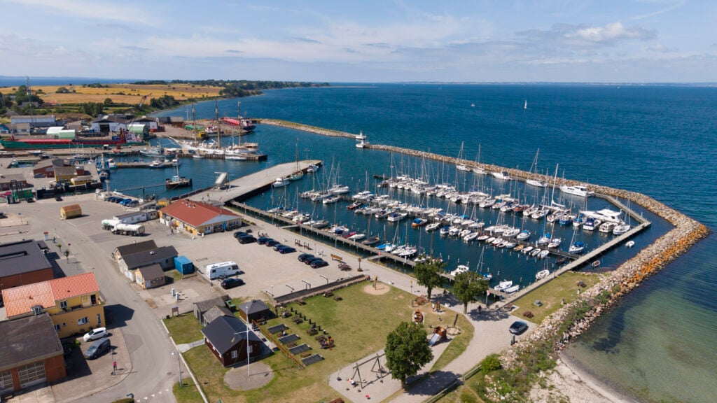 Luftaufnahme eines Hafens: Yachten und Segelboote hinter Steinmole, umliegende Werftgebäude, blaues Meer, klarer Himmel.