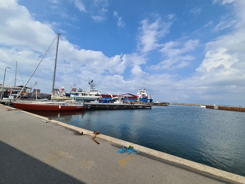 Ruhiger Hafen mit rotem Segelboot am Kai, mehreren größeren Schiffen im Hintergrund unter teils bewölktem Himmel.
