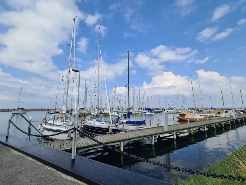 Bootshafen mit vielen festgemachten Segelbooten an einem Holzsteg, ruhiges Wasser unter blauem Himmel mit Wolken.