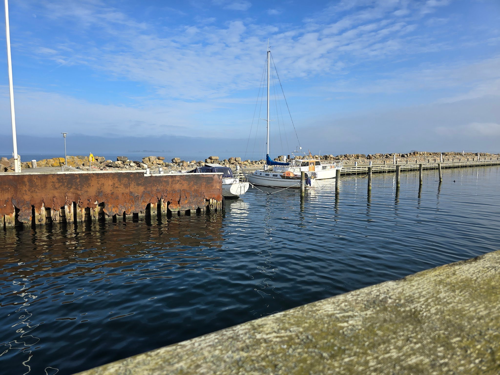 Kleiner Hafen; weißes Segelboot liegt an Pfahlpier, ruhiges Wasser, blauer Himmel, rostige Kaimauer links.