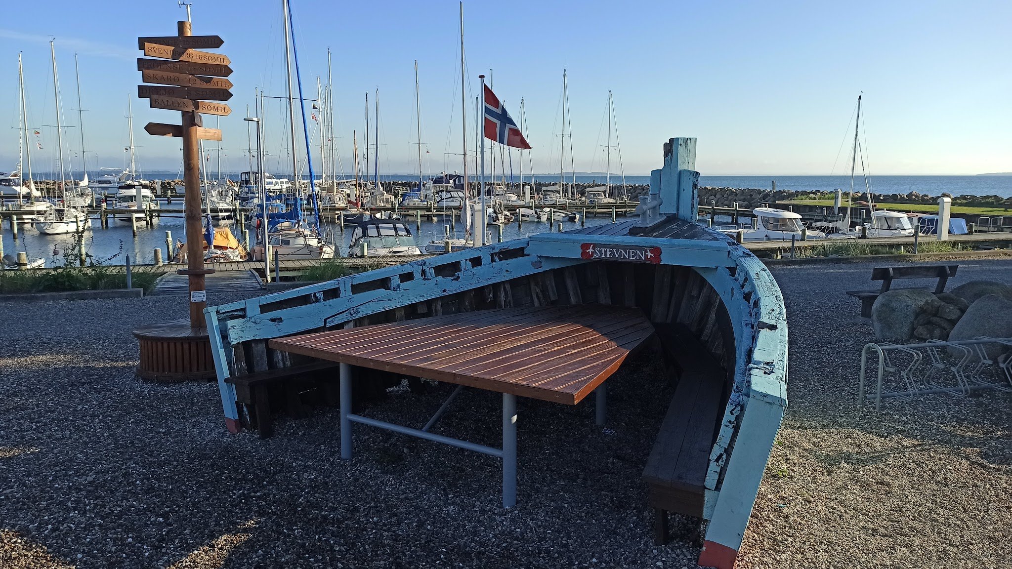 Altes türkis Boot als Picknicktisch vor Jachthafen; norwegische Flagge weht, daneben hölzerner Wegweiser