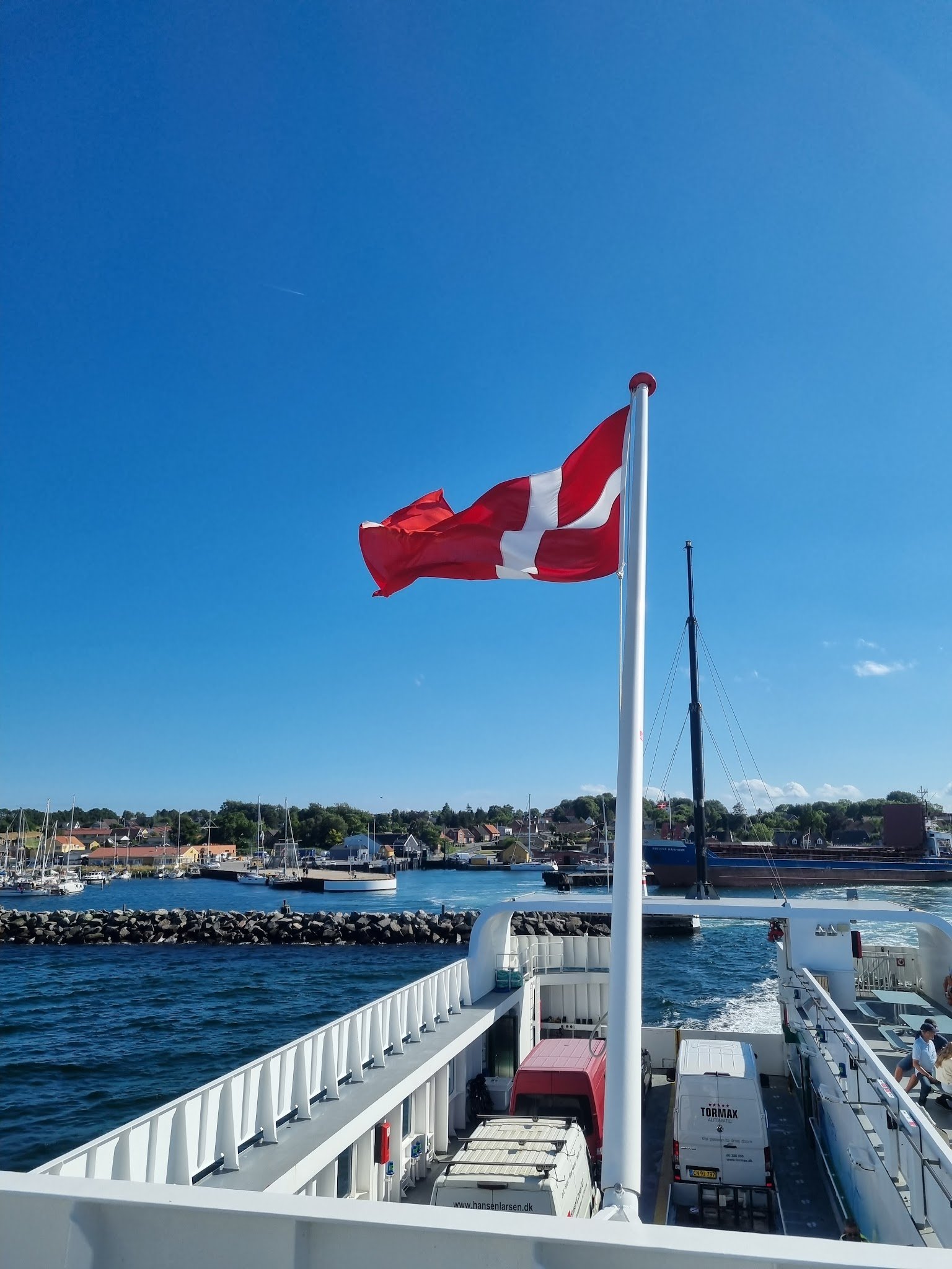 Dänische Flagge weht auf Fähre, darunter Fahrzeuge; im Hintergrund Hafen mit Segelbooten und Küstenhäusern, wolkenloser Himmel.