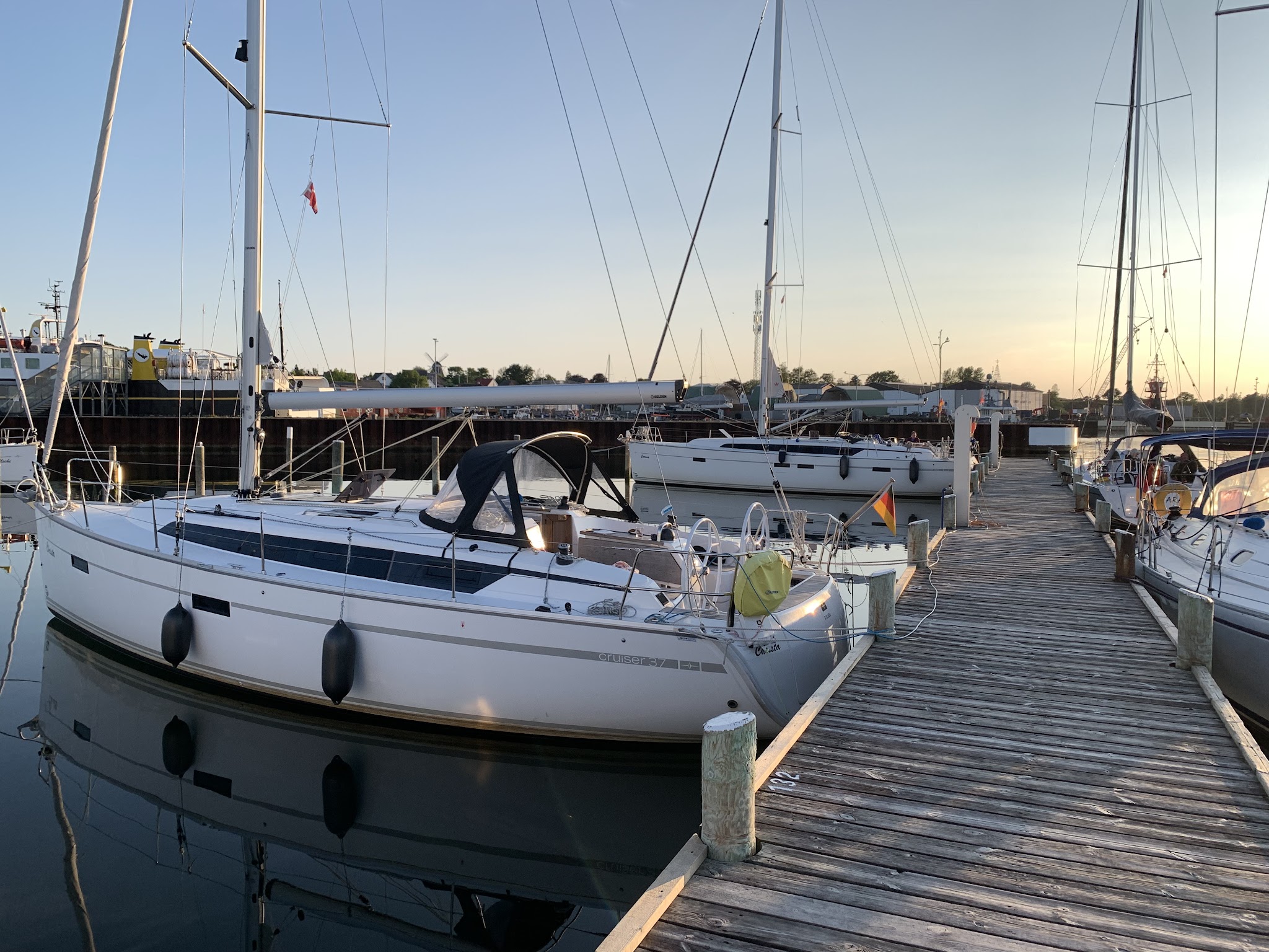 Segelboote vertäut an hölzernem Steg, ruhiges Hafenwasser spiegelt Masten im warmen Licht des Sonnenuntergangs.