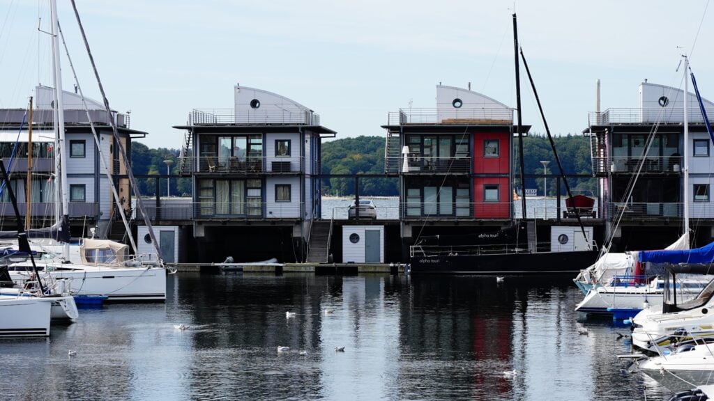Mehrstöckige Hausboote am Kai, umgeben von vertäuten Segeljachten; ruhiges Wasser mit Möwen.