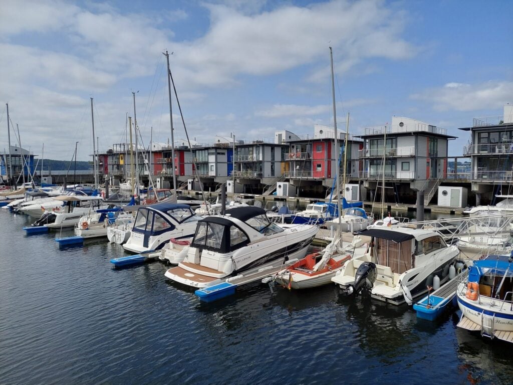 Mehrere Boote liegen im Jachthafen vor modernen, bunt gestrichenen Stelzenhäusern unter leicht bewölktem Himmel.
