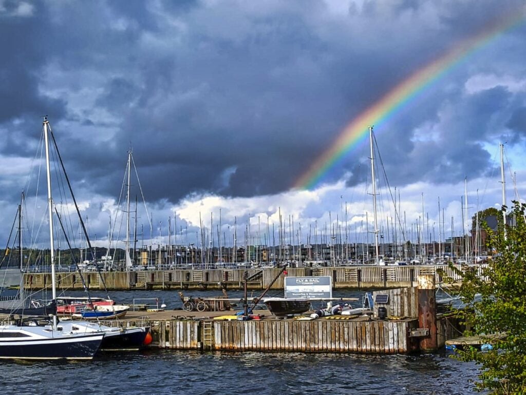 Bunter Regenbogen vor dunklen Wolken über Yachthafen mit vielen Segelbootmasten und leicht bewegtem Wasser.
