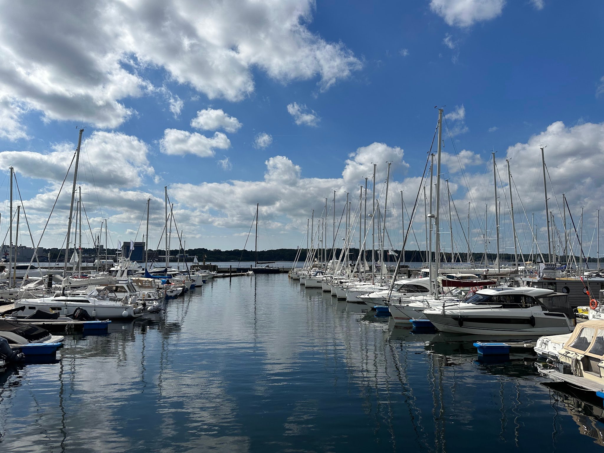 Ruhiger Yachthafen; Reihen weißer Segelboote spiegeln sich im Wasser unter blauem Himmel mit Wolken.