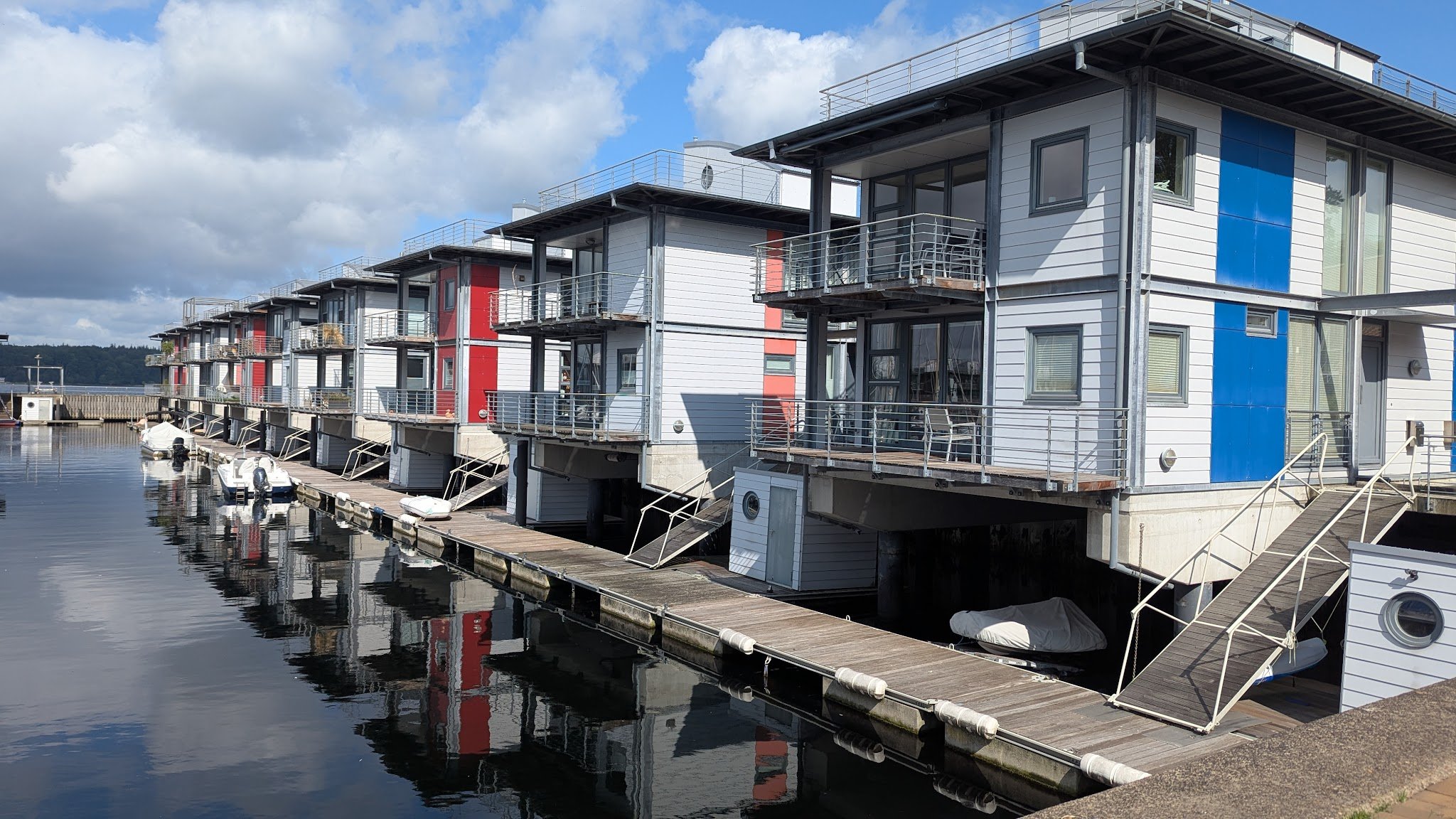 Reihe moderner Hausboote mit bunten Fassaden an Holzstegen, Boote spiegeln sich im ruhigen Wasser.